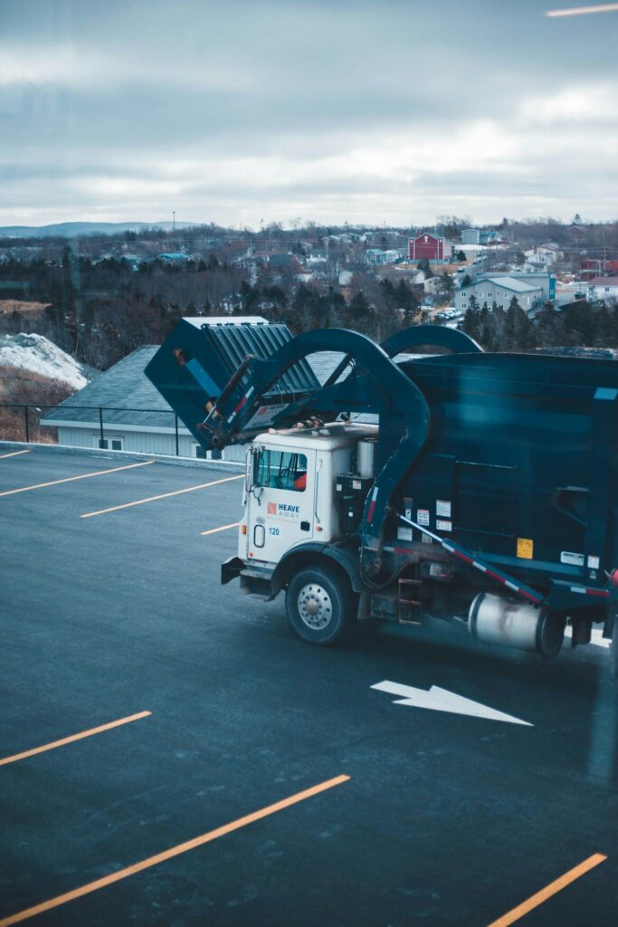 Garbage truck parked in an empty lot with a town in the background, under overcast skies.